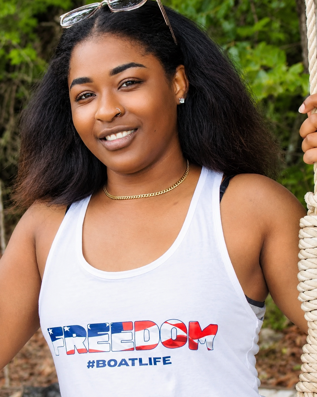A woman wearing a white tank top with 'FREEDOM #BOATLIFE' printed in red, white, and blue on the front, standing outdoors on a rope swing with green foliage in the background.