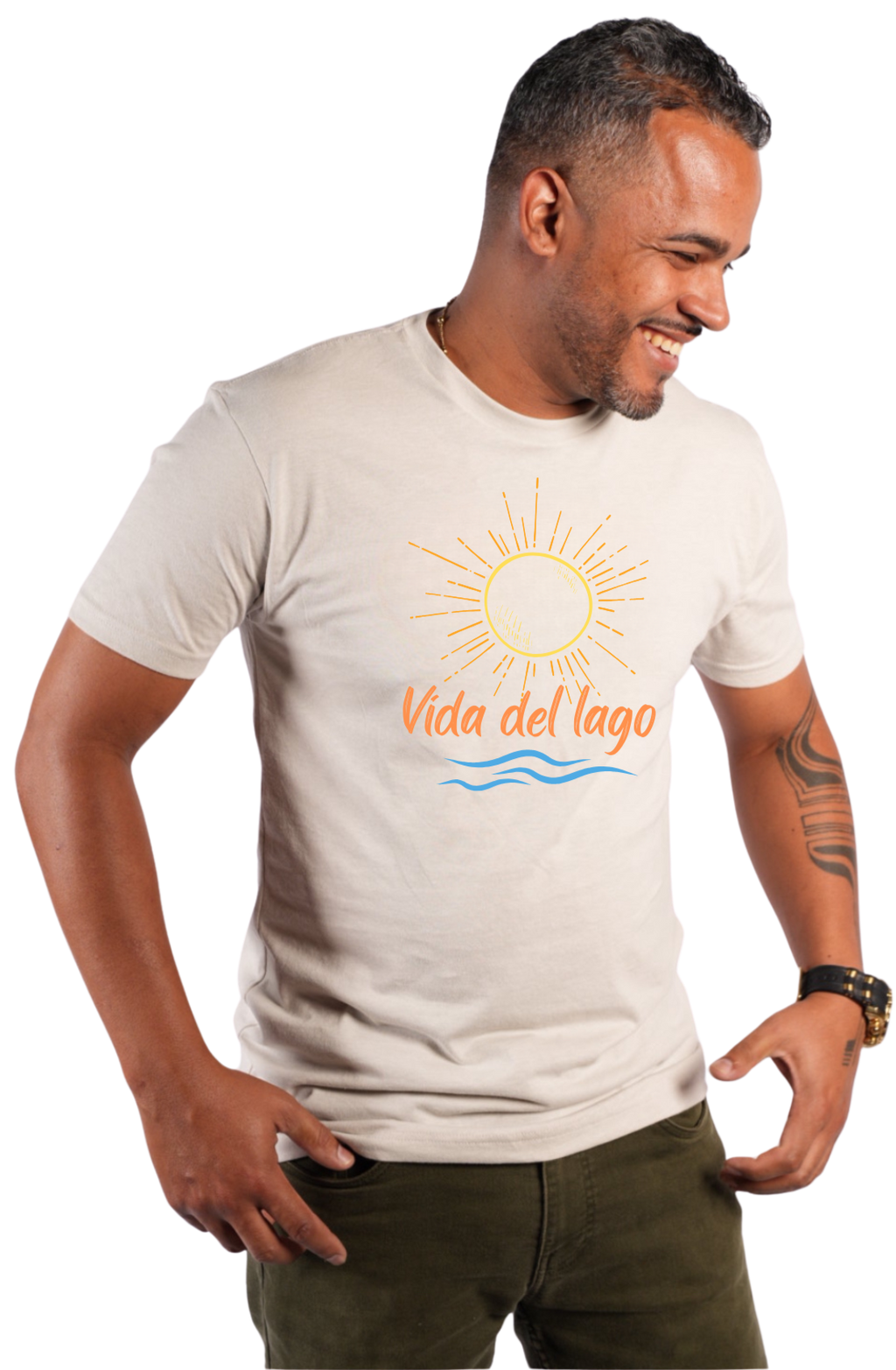 A man wearing a light beige crewneck t-shirt with a graphic print of a yellow sun, orange text reading 'Vida del lago', and blue waves. He is also wearing dark olive green pants, set against a plain white background.