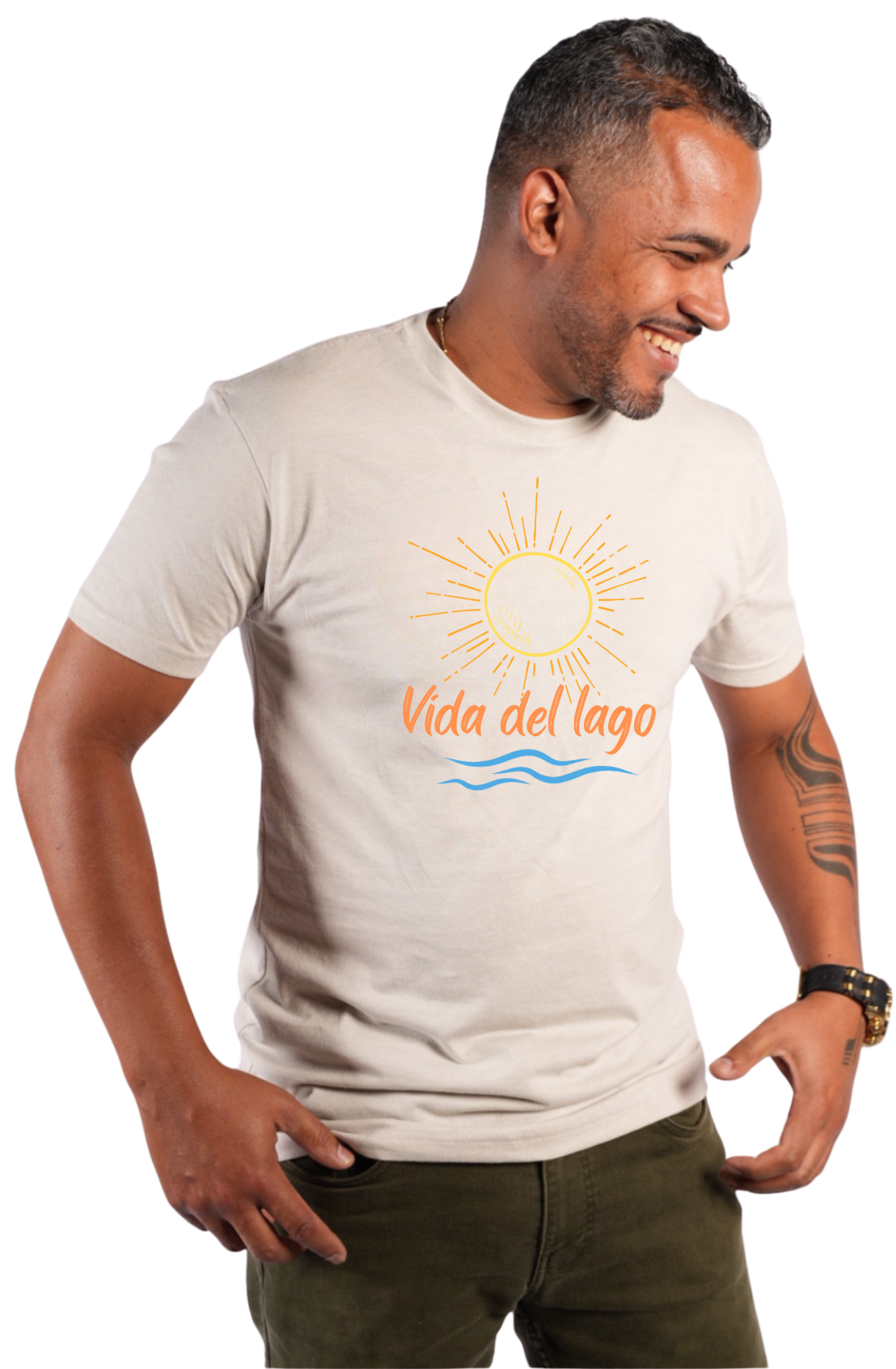 A man wearing a light beige crewneck t-shirt with a graphic print of a yellow sun, orange text reading 'Vida del lago', and blue waves. He is also wearing dark olive green pants, set against a plain white background.
