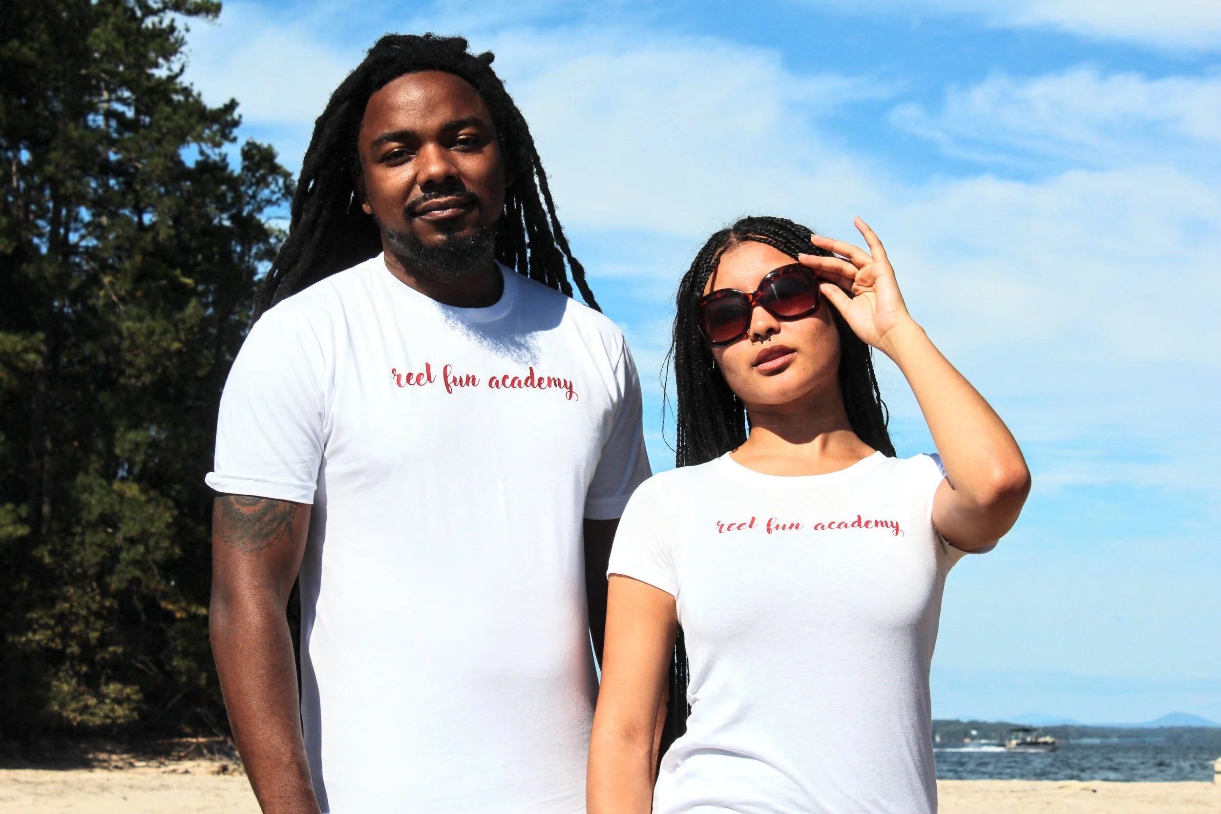 A man and a woman standing on a beach wearing matching white t-shirts with 'real fun academy' printed in red script across the chest.
