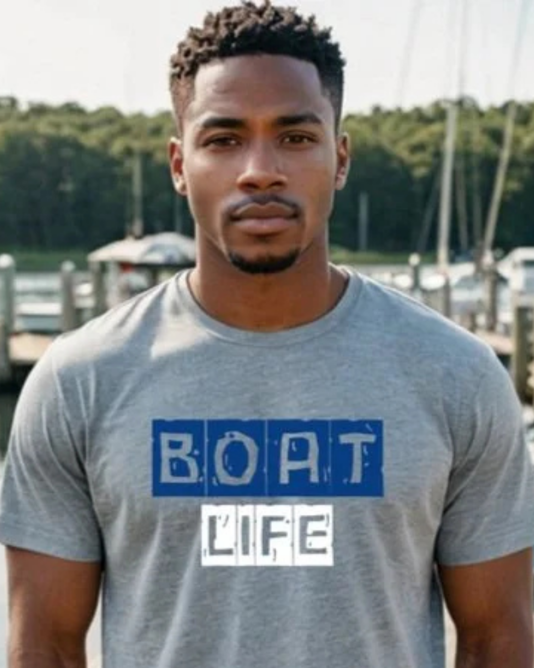 A man wearing a heather gray t-shirt with a graphic that reads 'BOAT LIFE' in blue and white block letters, standing outdoors in front of a blurred marina background.