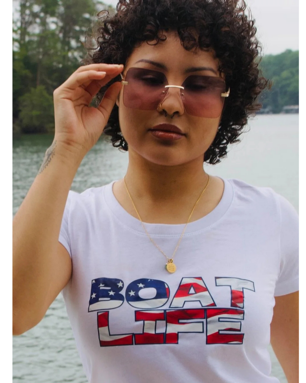 A woman with curly dark hair wearing a white graphic t-shirt with 'BOAT LIFE' printed in an American flag pattern, holding her sunglasses against a background of water and trees.
