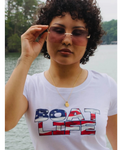 A woman with curly dark hair wearing a white graphic t-shirt with 'BOAT LIFE' printed in an American flag pattern, holding her sunglasses against a background of water and trees.