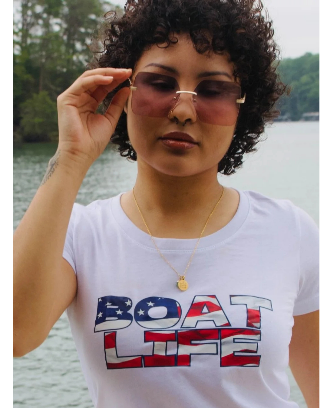 A woman with curly dark hair wearing a white graphic t-shirt with 'BOAT LIFE' printed in an American flag pattern, holding her sunglasses against a background of water and trees.