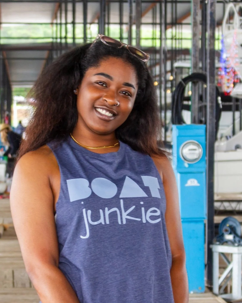 A smiling woman with curly hair wearing a heathered navy blue tank top with 'boat junkie' printed in white across the chest, at a boat marina.