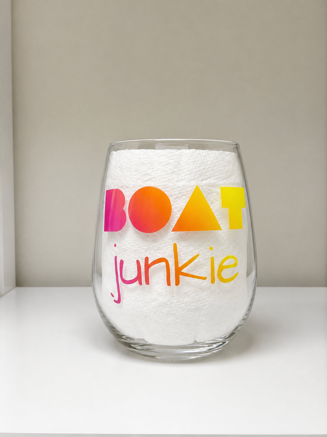 A clear shatterproof stemless wine glass with the words 'BOAT junkie' printed on it in colorful gradient lettering, set against a neutral beige and white background.


