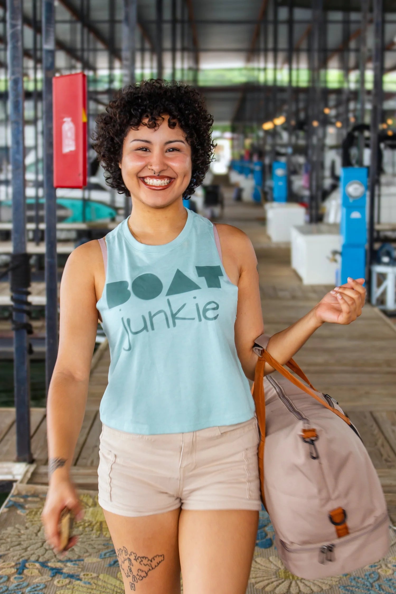 A smiling woman with curly dark hair walking down a boat dock wearing a light green  tank top with 'BOAT junkie' printed in dark teal, paired with beige shorts and carrying a large beige weekend bag.