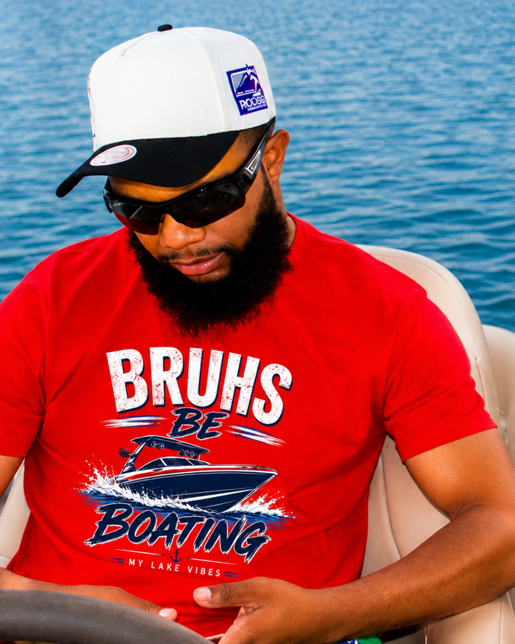 A man wearing a red graphic t-shirt with 'BRUHS BE BOATING MY LAKE VIBES' printed on the front, along with a white and black baseball cap and sunglasses, sitting on a boat with blue water in the background.