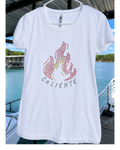 A white short-sleeve t-shirt hanging on a white hanger, featuring a rhinestone flame graphic in red, pink, and yellow with the word 'CALIENTE' in silver rhinestones below it. The background shows a sunny outdoor scene with a blue swimming pool and green trees.
