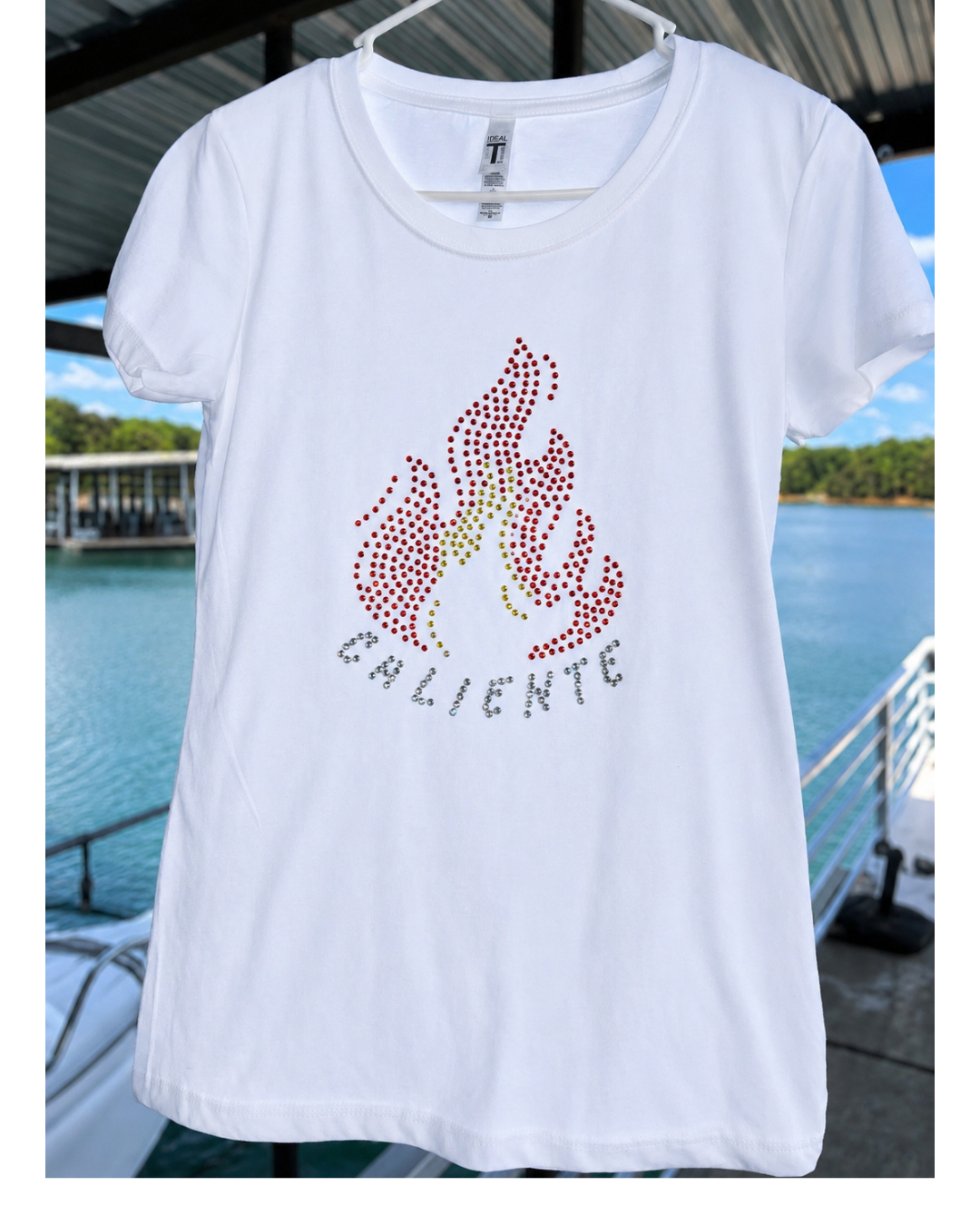 A white short-sleeve t-shirt hanging on a white hanger, featuring a rhinestone flame graphic in red, pink, and yellow with the word 'CALIENTE' in silver rhinestones below it. The background shows a sunny outdoor scene with a blue swimming pool and green trees.