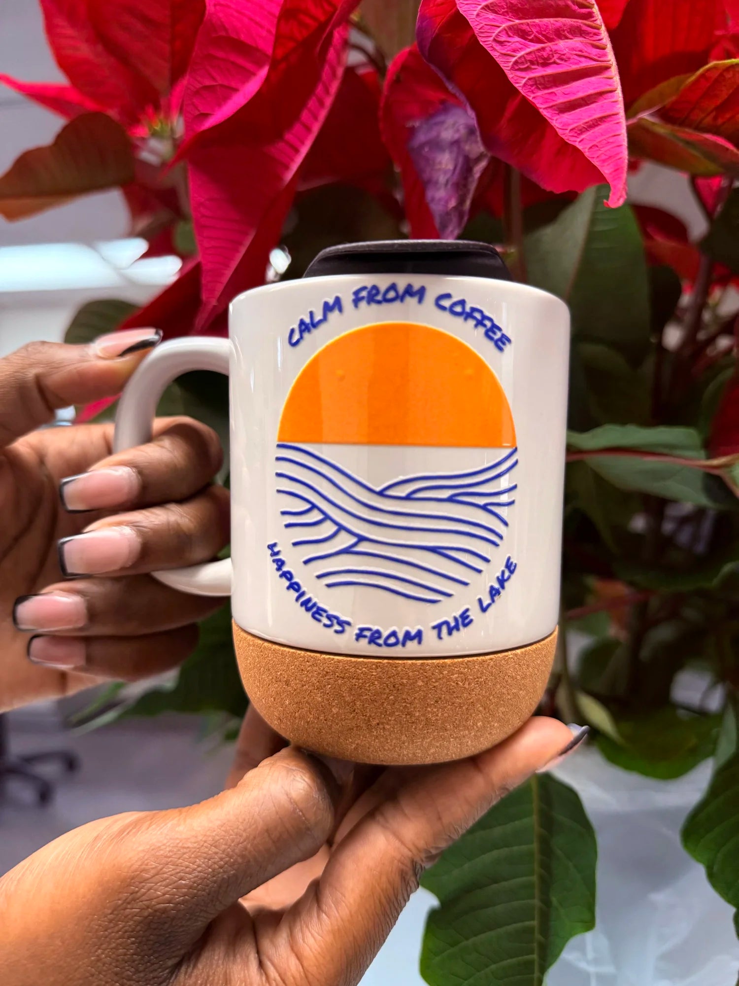 A ceramic coffee mug with a white upper body, orange and blue graphic design, and a natural cork base, held by two hands with wording "CALM FROM COFFEE...HAPPINESS FROM THE LAKE"