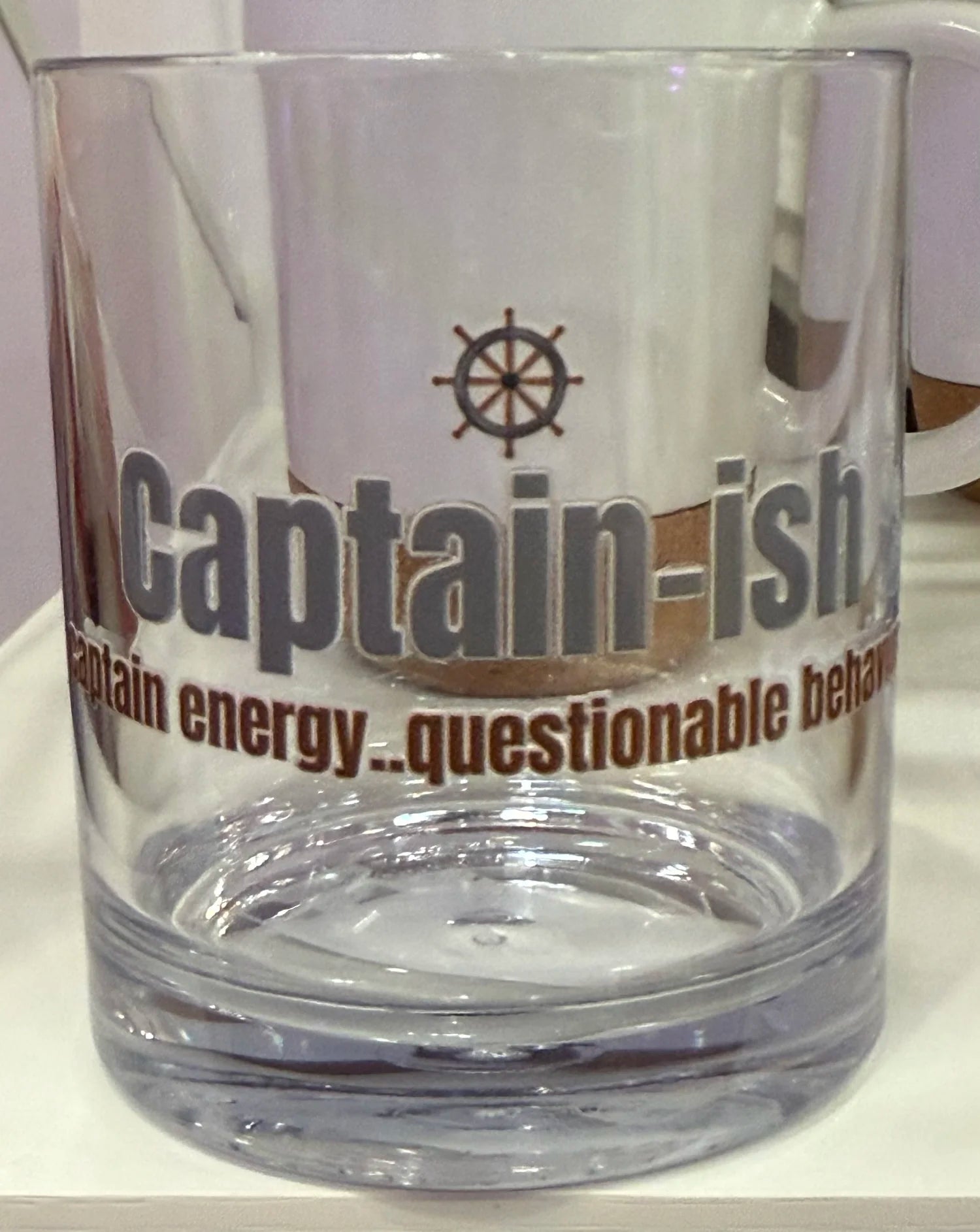 A clear glass tumbler with a thick base, featuring a dark brown ship's wheel graphic and the text 'Captain-ish' and 'Captain energy.