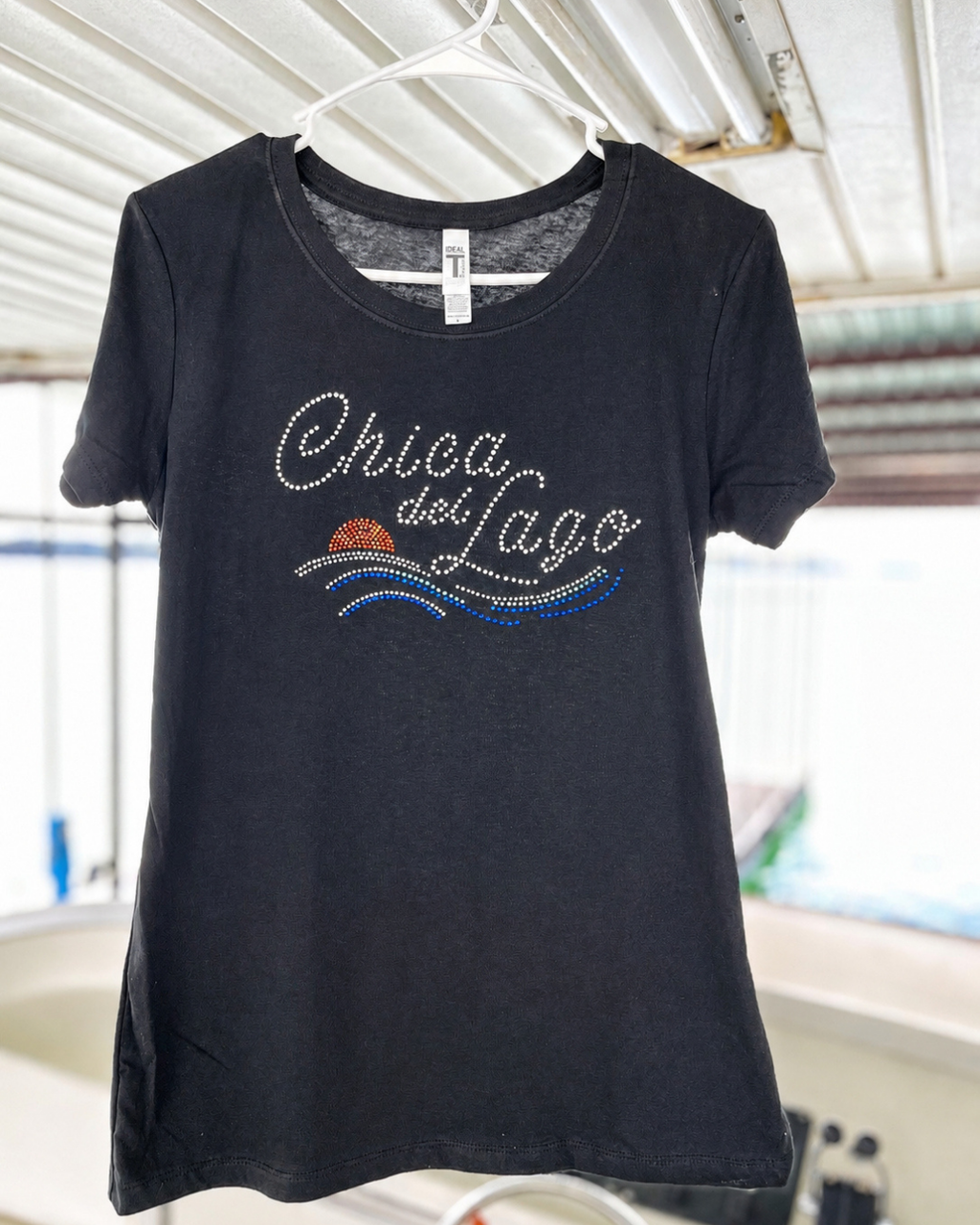 A black short-sleeve t-shirt hanging on a white plastic hanger, featuring a rhinestone-embellished graphic that reads 'Chica del Lago' with blue and orange wave details. The shirt is photographed in a casual setting with a corrugated metal roof visible in the background.