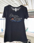 A black short-sleeve t-shirt hanging on a white plastic hanger, featuring a rhinestone-embellished graphic that reads 'Chica del Lago' with blue and orange wave details. The shirt is photographed in a casual setting with a corrugated metal roof visible in the background.
