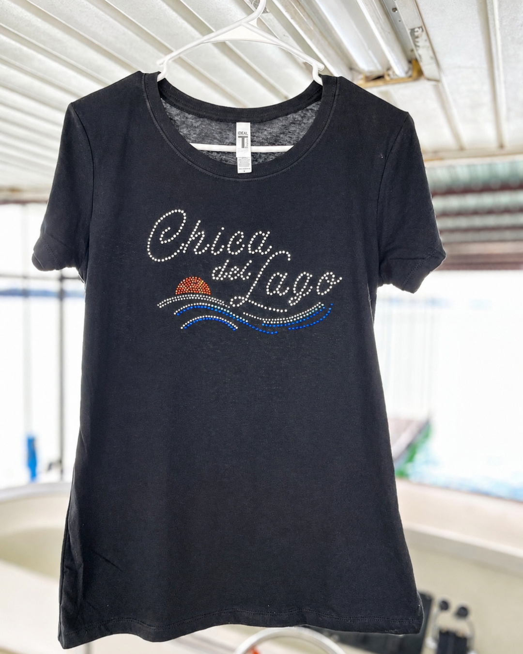 A black short-sleeve t-shirt hanging on a white plastic hanger, featuring a rhinestone-embellished graphic that reads 'Chica del Lago' with blue and orange wave details. The shirt is photographed in a casual setting with a corrugated metal roof visible in the background.