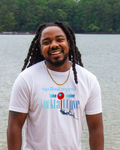 A  smiling man with long black locks stands outdoors in front of a body of water and trees, wearing a white graphic T-shirt with blue and red text reading 'sip, float, repeat' and 'Lake Tanner Cocktail Cove,' along with a small red-and-blue pin of the location.