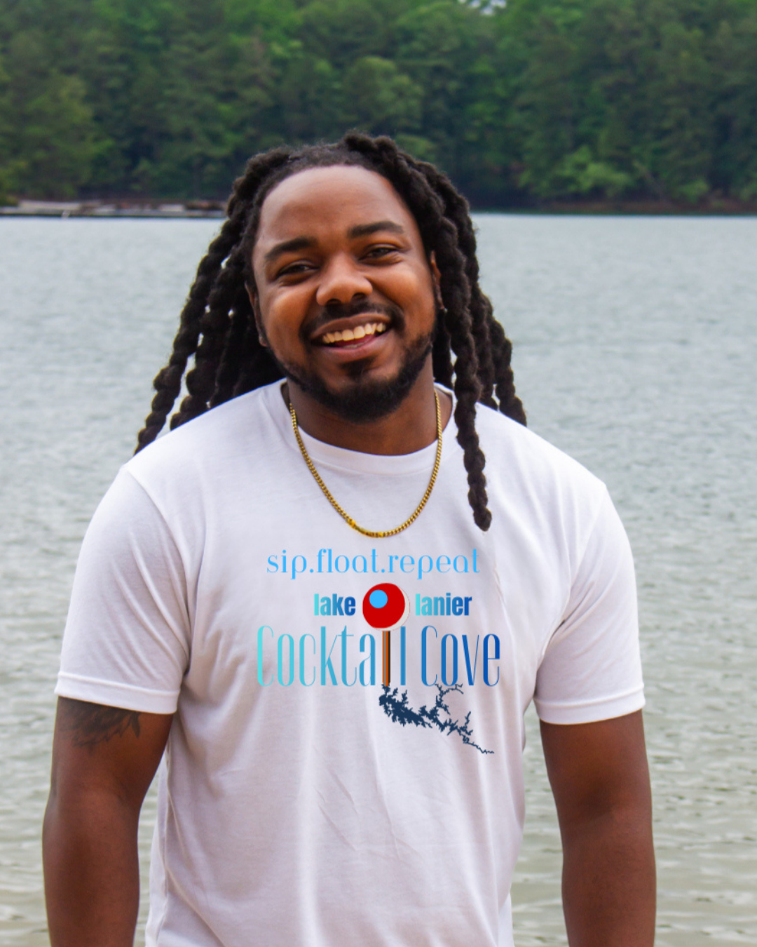A  smiling man with long black locks stands outdoors in front of a body of water and trees, wearing a white graphic T-shirt with blue and red text reading 'sip, float, repeat' and 'Lake Tanner Cocktail Cove,' along with a small red-and-blue pin of the location.