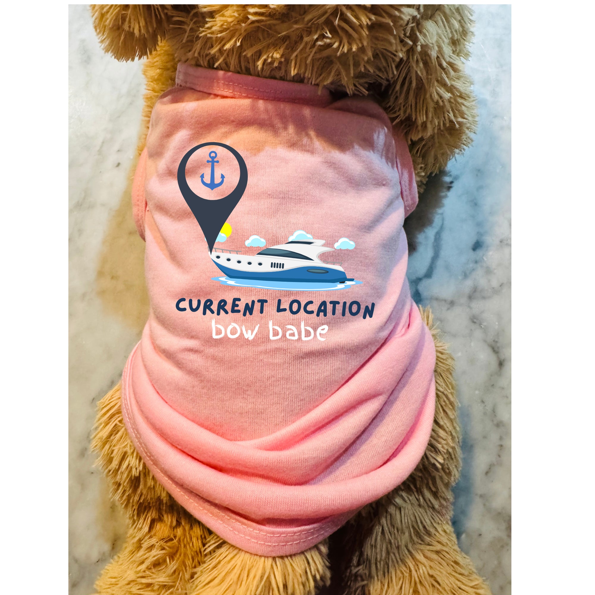 A pink pet shirt worn by a fluffy brown dog, featuring a graphic of a boat under a location pin with the text 'CURRENT LOCATION bow babe'.