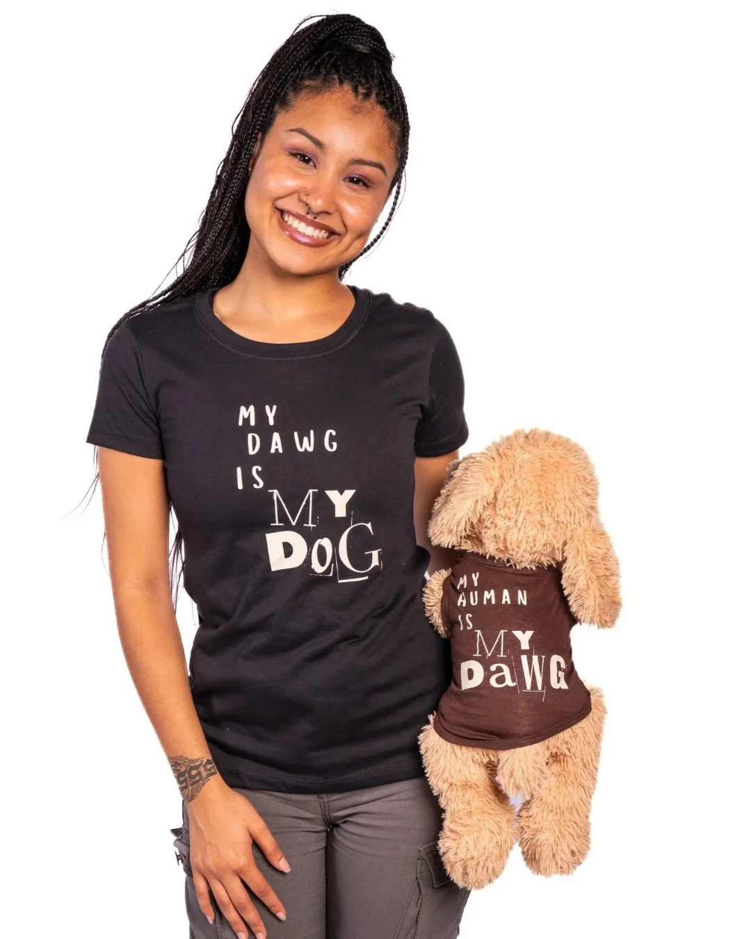 A woman wearing a black graphic t-shirt that reads 'MY DAWG IS MY DOG' holds a plush tan dog toy wearing a brown shirt that reads 'MY HUMAN IS MY DAWG' against a plain white background.