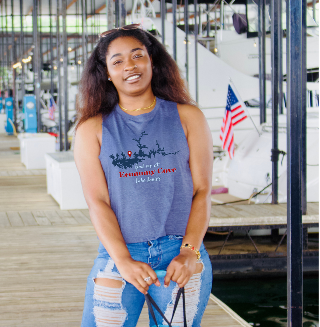 A woman wearing a blue cropped sleeveless tank top with a blue and red graphic print that reads 'find me at Economy Cove Lake Lanier, posed on a boat dock
