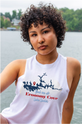 A woman with curly dark hair wearing a white sleeveless tank top with a blue and red graphic print that reads 'find me at Economy Cove Lake Lanier, posed outdoors with water and trees in the background.