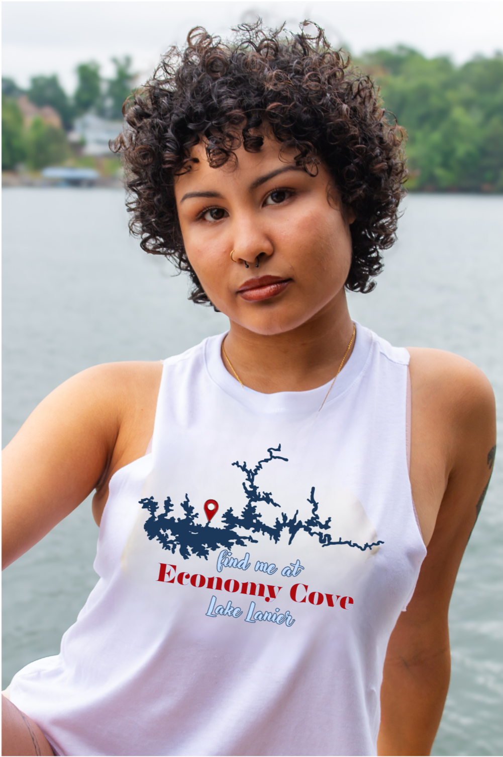 A woman with curly dark hair wearing a white sleeveless tank top with a blue and red graphic print that reads 'find me at Economy Cove Lake Lanier, posed outdoors with water and trees in the background.