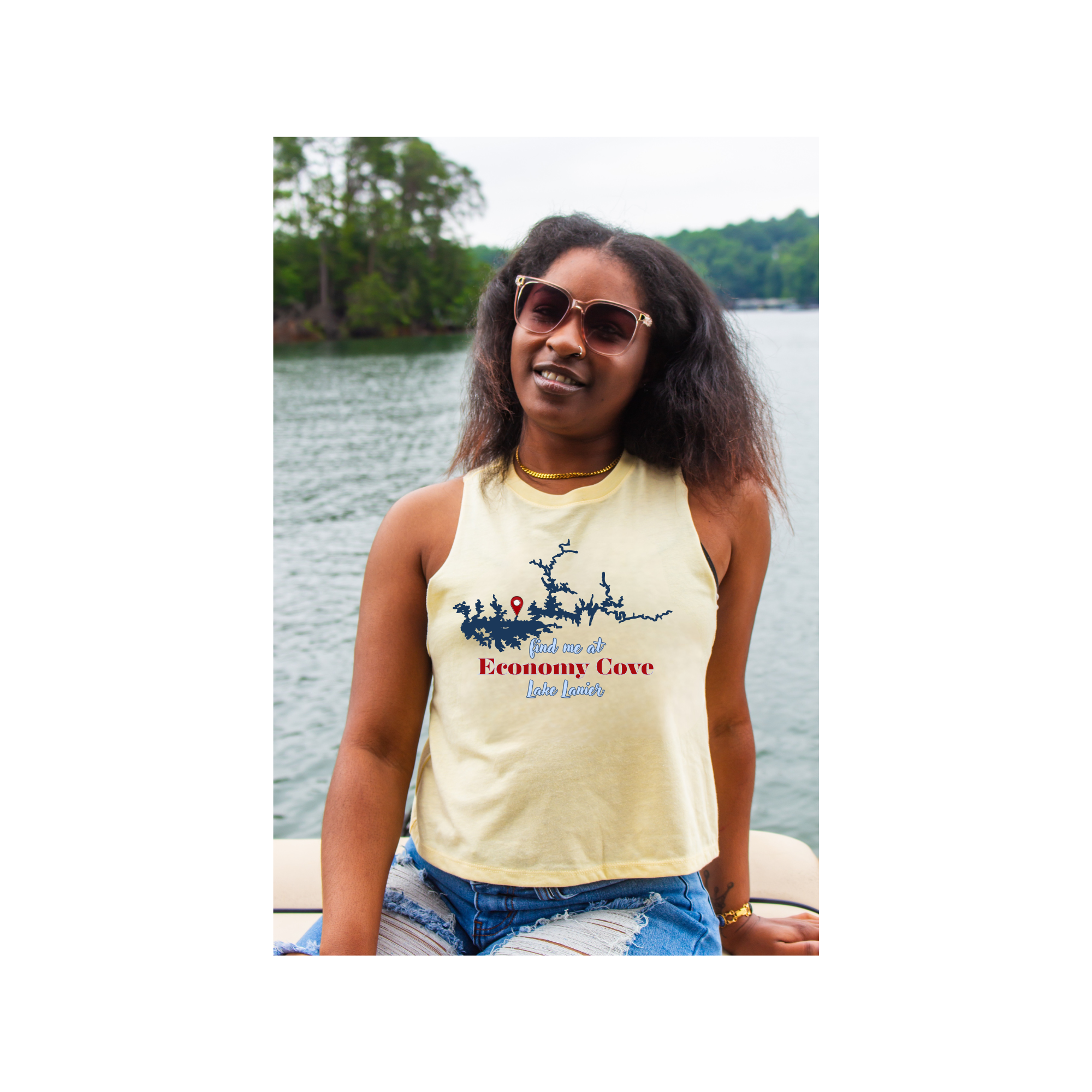 A woman wearing a pale yellow sleeveless tank top with a blue and red 'Economy Cove' graphic, sitting on a boat with water and trees in the background.