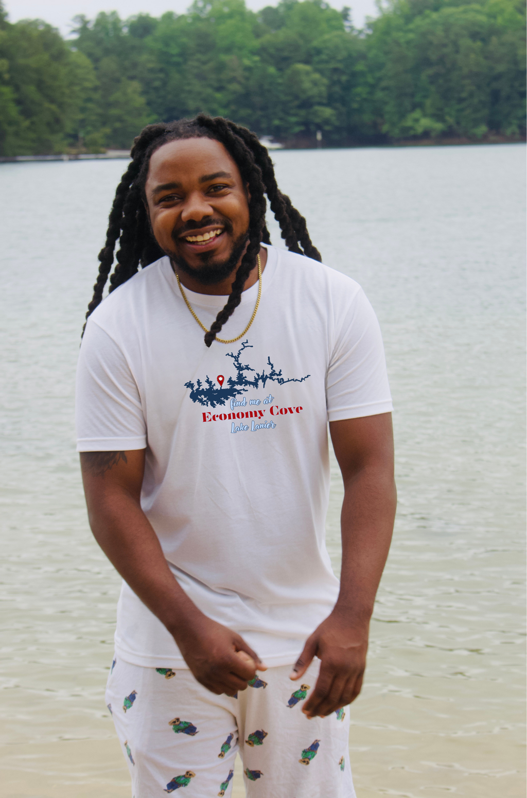 A smiling man with long black locks stands outdoors by the water wearing a white graphic T-shirt and white shorts with small green fish prints. The shirt has a blue graphic of LAKE LANIER sand red/blue text reading 'Economy Cove'.