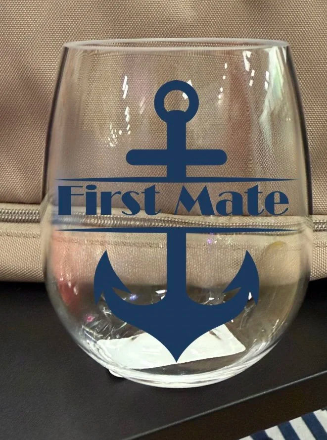 A clear shatterproof stemless wine glass with a navy blue anchor graphic and the words 'First Mate' printed on the front, shown on a dark surface with a beige textured bag in the background.