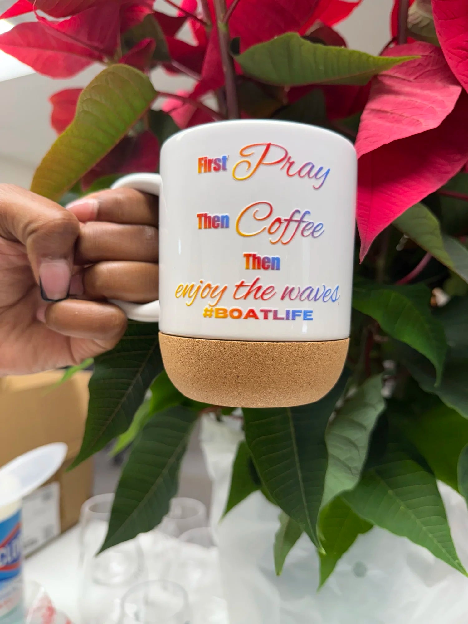 A hand holds a white ceramic coffee mug with a cork base. The mug features the text 'First Pray Then Coffee Then enjoy the waves #BOATLIFE' in colorful script.
