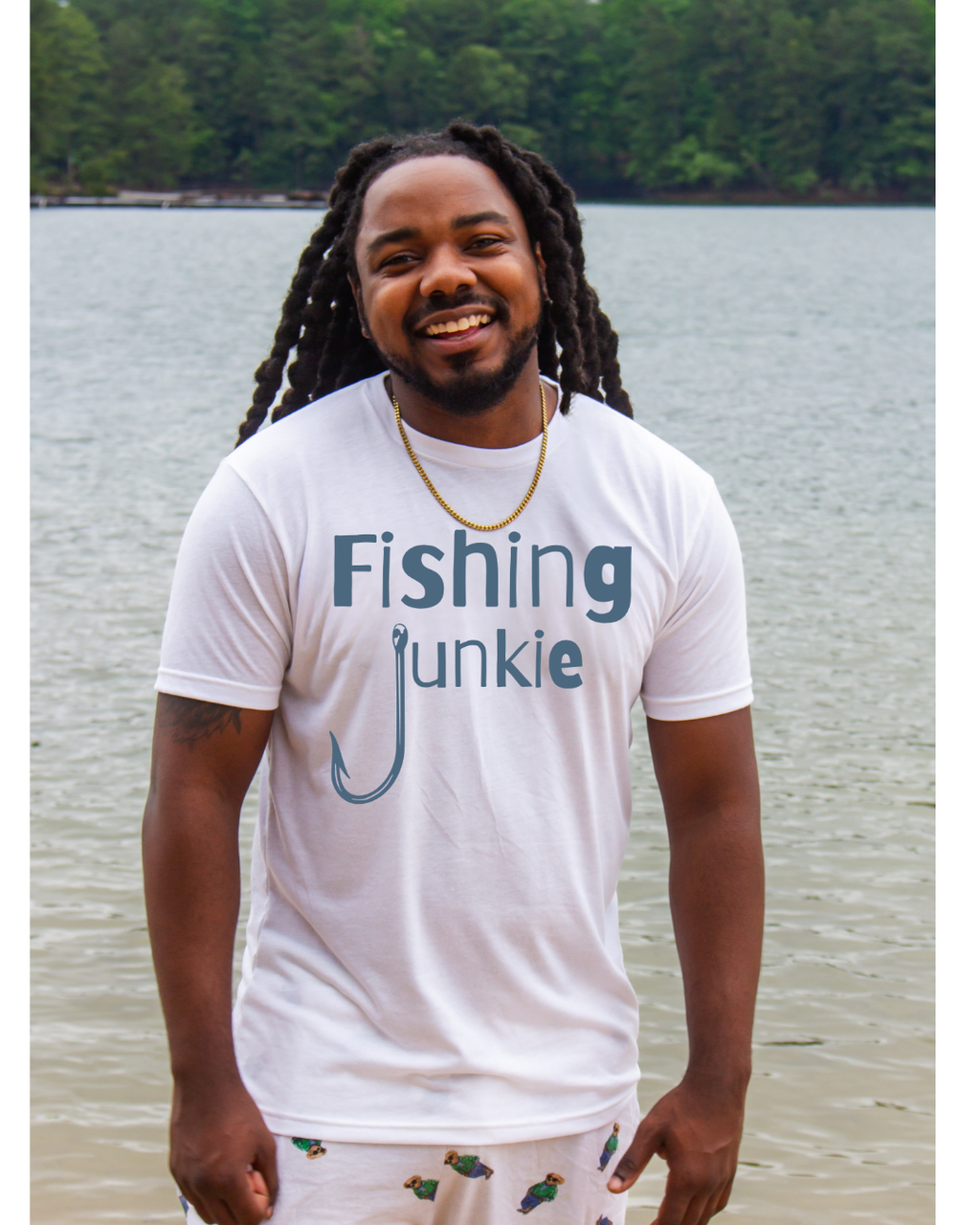 A smiling man with long dreadlocks stands outdoors by the water wearing a white graphic T-shirt that reads 'Fishing Junkie' in blue-gray lettering with a fishing hook graphic, plus white shorts with small fish prints.