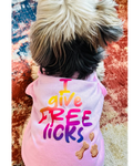 A small fluffy dog seen from behind wearing a pink pet shirt with the text 'I give FREE licks' in a rainbow gradient and small bone and paw prints on the back, resting on a patterned rug.
