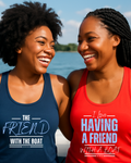 Two smiling women standing outdoors by the water, each wearing a racerback tank top with a slogan. The woman on the left wears a navy tank top that says 'THE FRIEND WITH THE BOAT,' and the woman on the right wears a red tank top that says 'I Love HAVING A FRIEND WITH A BOAT.