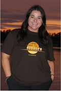 A smiling woman with long dark hair stands outdoors at sunset wearing a dark brown graphic T-shirt with a yellow and orange circular sun design and the text 'HERE FOR THE SUNSETS' and '#LAKELIFE'.

