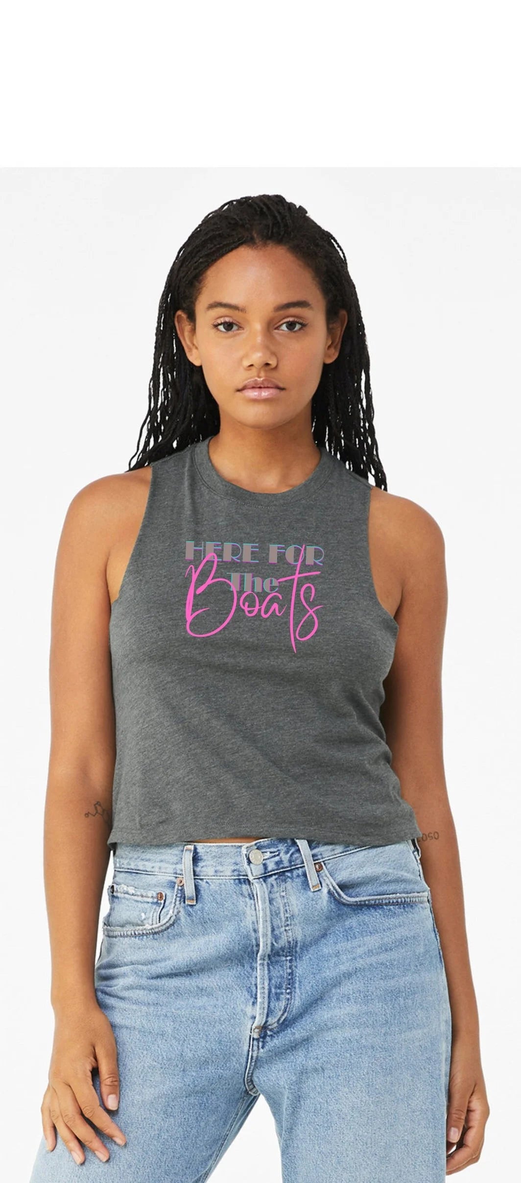 A woman modeling a dark gray sleeveless crop top with pink and white text that reads 'HERE FOR THE BOATS', paired with light blue jeans, against a plain white background.