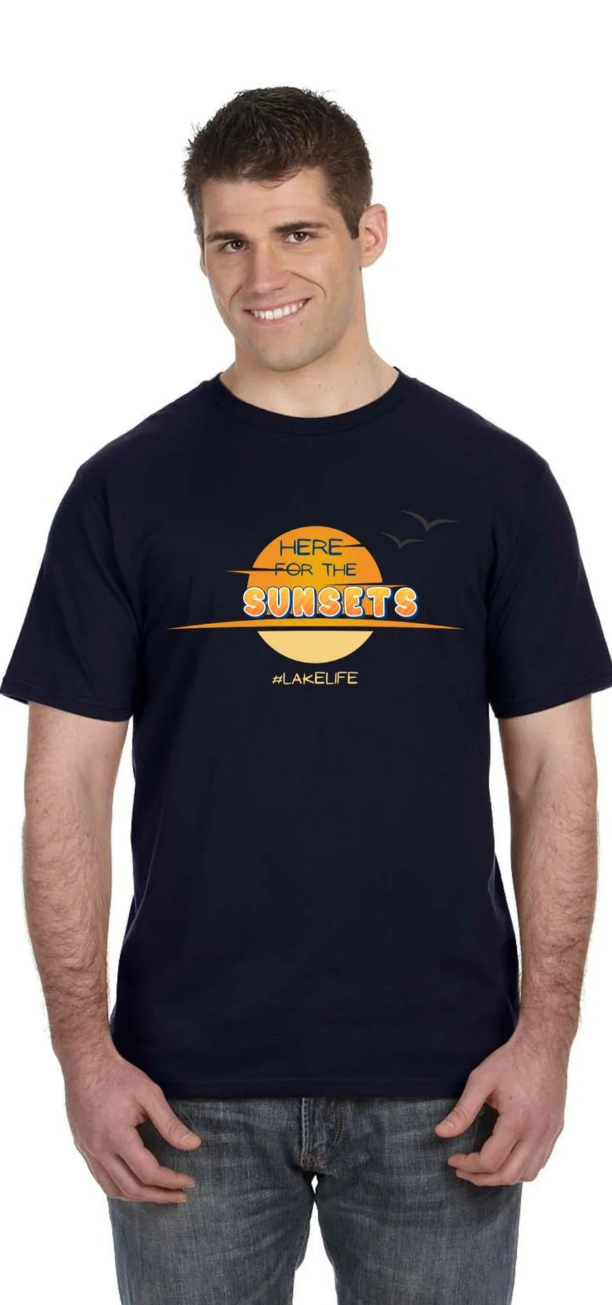 A man wearing a navy blue t-shirt with a retro-style orange and yellow graphic that reads 'HERE FOR THE SUNSETS' and '#LAKELIFE' underneath, paired with gray jeans, against a plain white background.