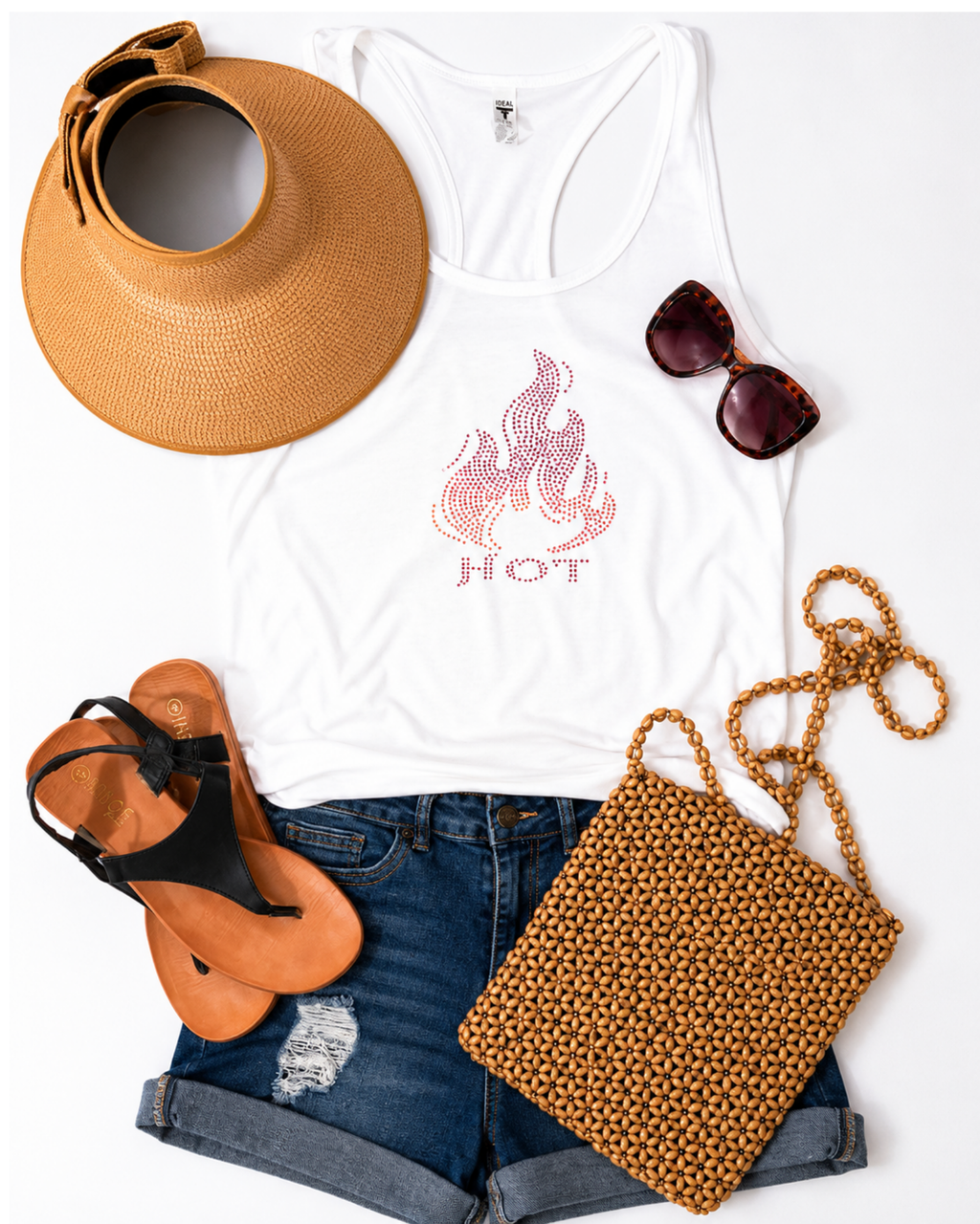 A styled flat-lay of a white racerback tank top with a pink flame graphic and the word 'HOT' on the front, arranged with a tan sun hat, dark sunglasses, blue denim shorts, tan sandals, and a tan woven shoulder bag on a white background.