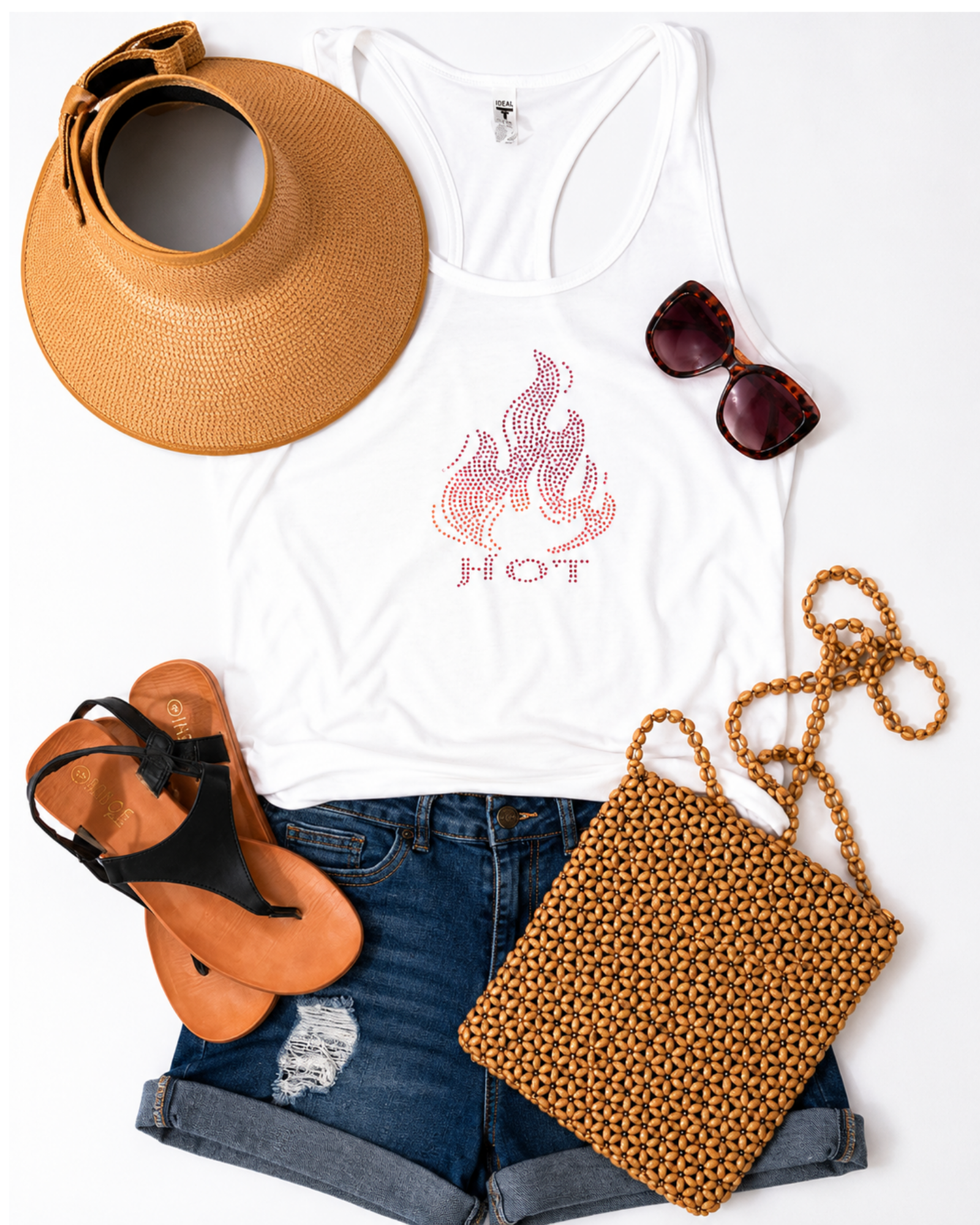 A styled flat-lay of a white racerback tank top with a pink flame graphic and the word 'HOT' on the front, arranged with a tan sun hat, dark sunglasses, blue denim shorts, tan sandals, and a tan woven shoulder bag on a white background.