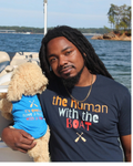 A man with long black dreadlocks stands on a boat holding a plush dog toy wearing a blue shirt. He is wearing a navy graphic t-shirt that reads 'the human With the BOAT' in orange, white, and red lettering and dog shirt says "it's good to have a human with a boat"