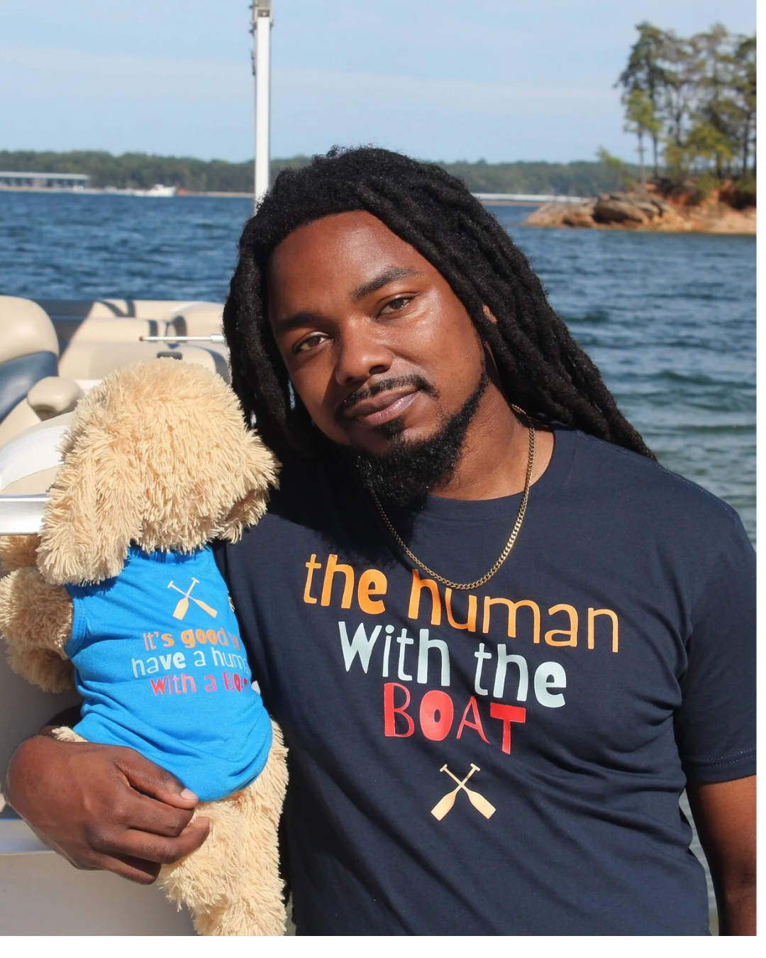 A man with long black dreadlocks stands on a boat holding a plush dog toy wearing a blue shirt. He is wearing a navy graphic t-shirt that reads 'the human With the BOAT' in orange, white, and red lettering and dog shirt says "it's good to have a human with a boat"