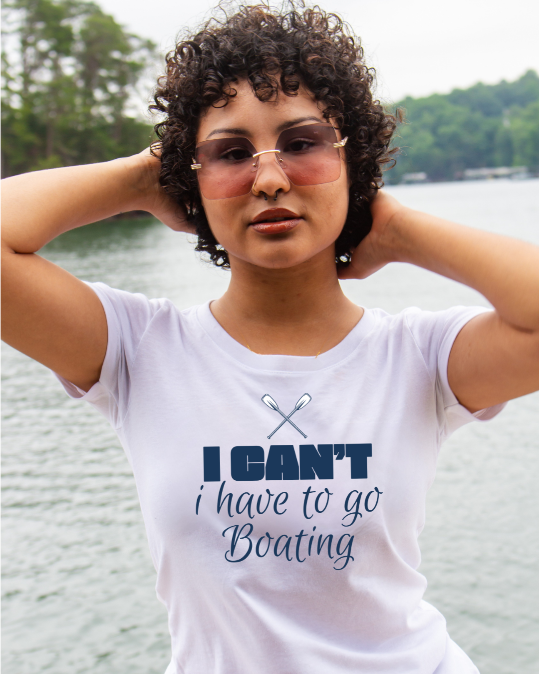 A woman with curly dark hair and pink-tinted sunglasses stands outdoors by the water wearing a white graphic T-shirt that reads 'I CAN'T i have to go Boating' with crossed oars printed above the text.