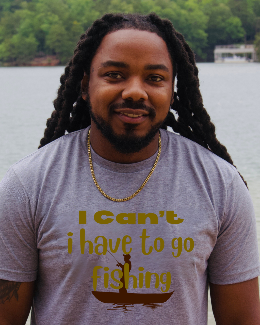 A man with long black dreadlocks stands outdoors by the water wearing a heather gray t-shirt with a gold graphic that reads 'I Can't i have to go fishing' above a fishing rod and boat illustration.