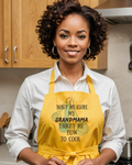 A smiling woman with curly dark hair stands in a kitchen wearing a white button-up shirt and a bright yellow apron with the printed phrase, 'I DON'T MEASURE, MY GRANDMAMA TAUGHT ME HOW TO COOK.'.