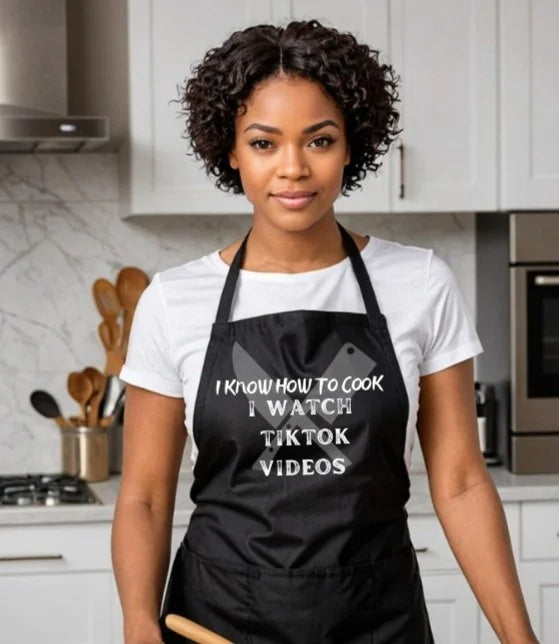 A woman with curly dark hair stands in a modern kitchen wearing a black apron over a white t-shirt. The apron features white text that reads 'I KNOW HOW TO COOK I WATCH TIKTOK VIDEOS' with a small white character graphic above the text.