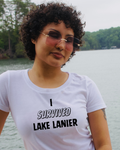 A woman with curly dark hair and pink-tinted sunglasses wearing a white graphic t-shirt that reads 'I SURVIVED LAKE LANIER' in black text, posed outdoors with a lake and trees in the background.