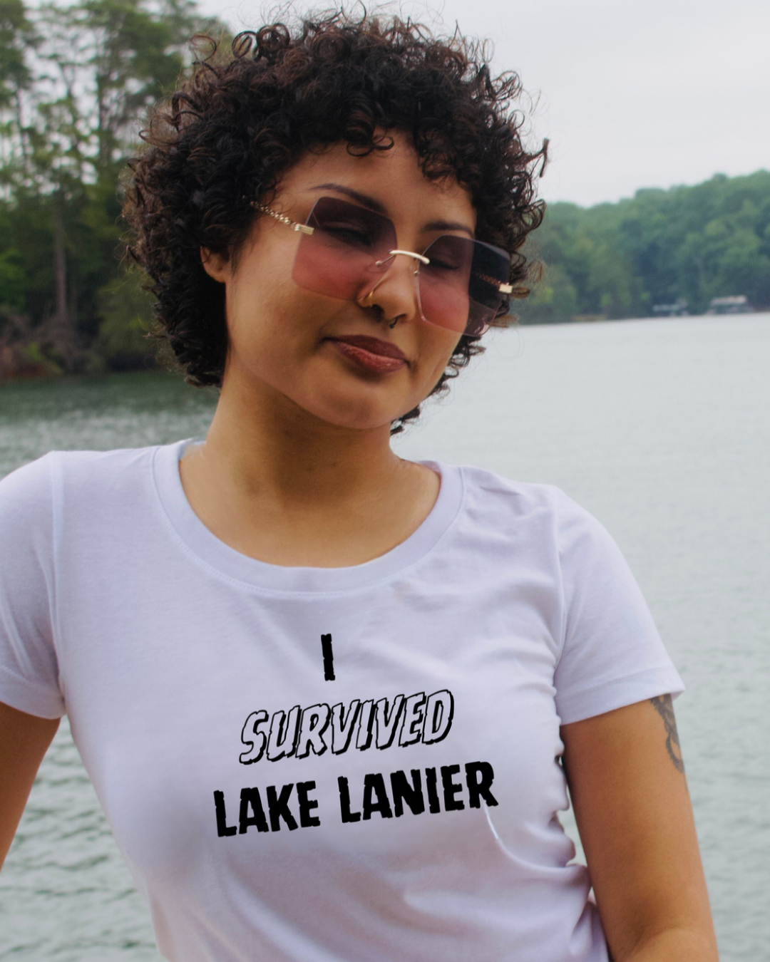 A woman with curly dark hair and pink-tinted sunglasses wearing a white graphic t-shirt that reads 'I SURVIVED LAKE LANIER' in black text, posed outdoors with a lake and trees in the background.