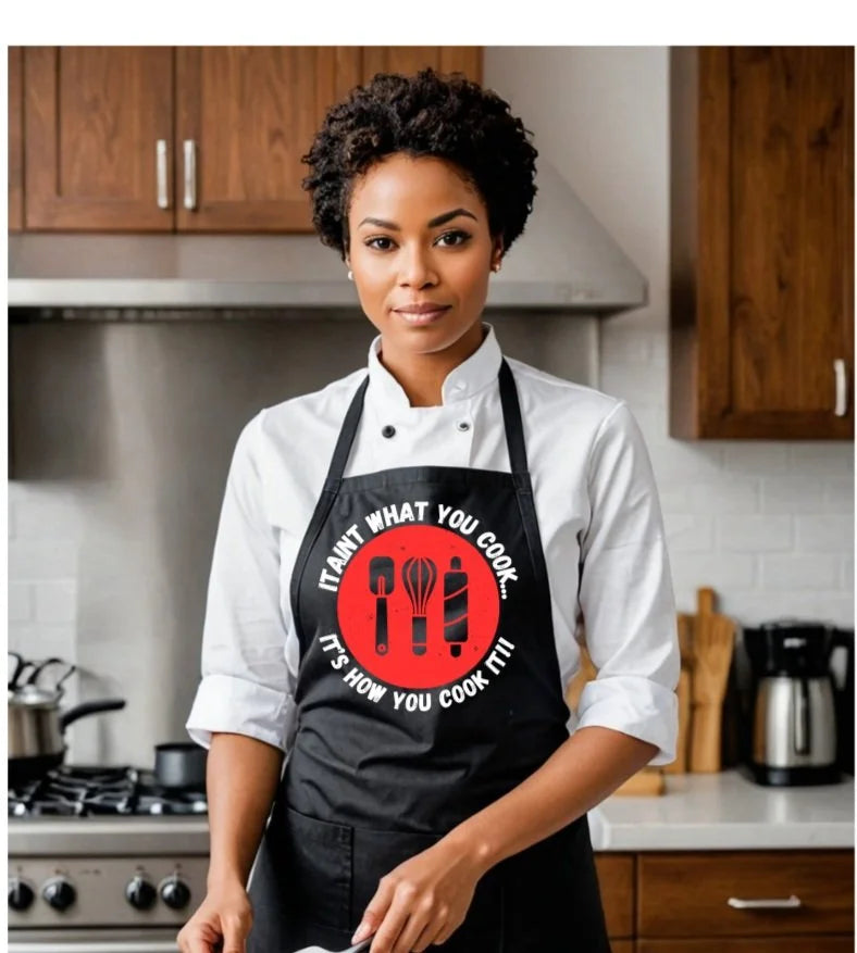 A woman in a kitchen wearing a white chef's coat and a black apron with a red circular graphic that reads 'IT AIN'T WHAT YOU COOK. IT'S HOW YOU COOK IT!'.
