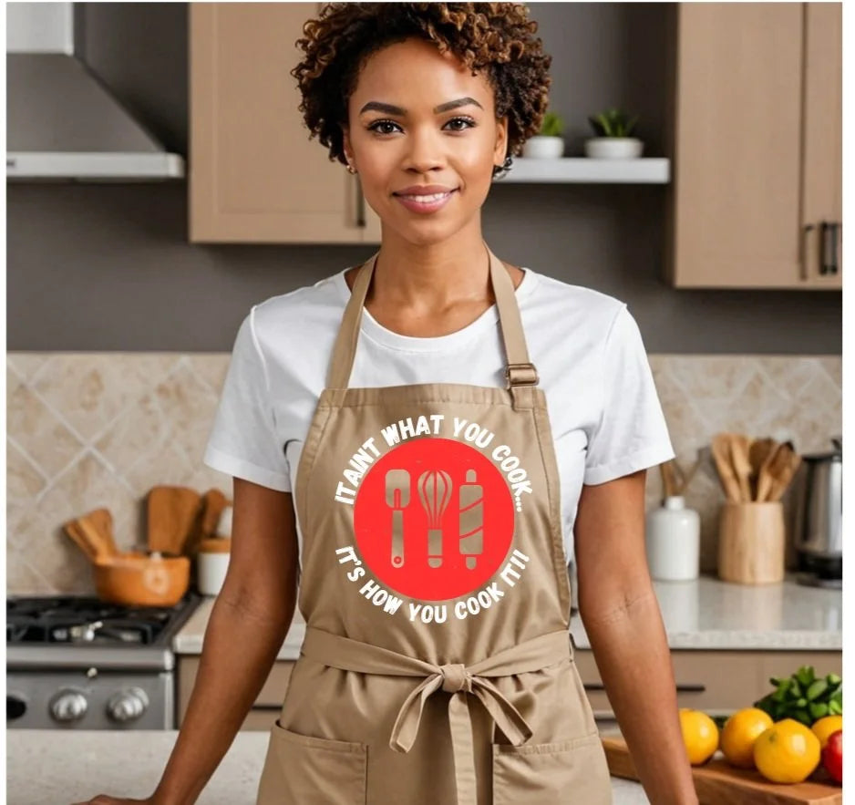 A woman wearing a beige apron with a red circular graphic that reads 'IT AIN'T WHAT YOU COOK, IT'S HOW YOU COOK IT!' stands in a kitchen setting.