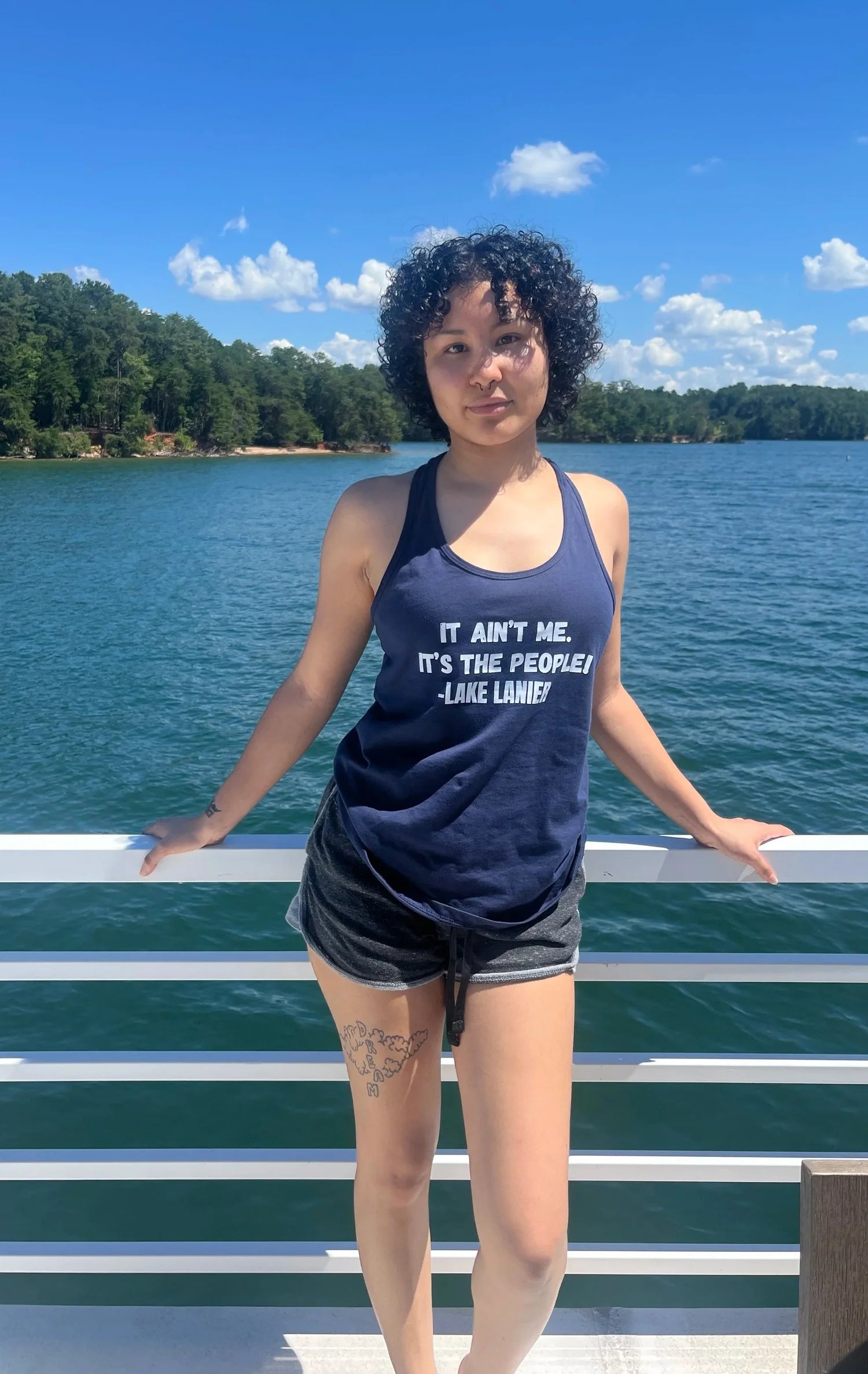 A woman stands on a boat deck with a lake and forest in the background, wearing a navy blue tank top with the white printed text 'IT AIN'T ME, IT'S THE PEOPLE! -LAKE LANE!'.
