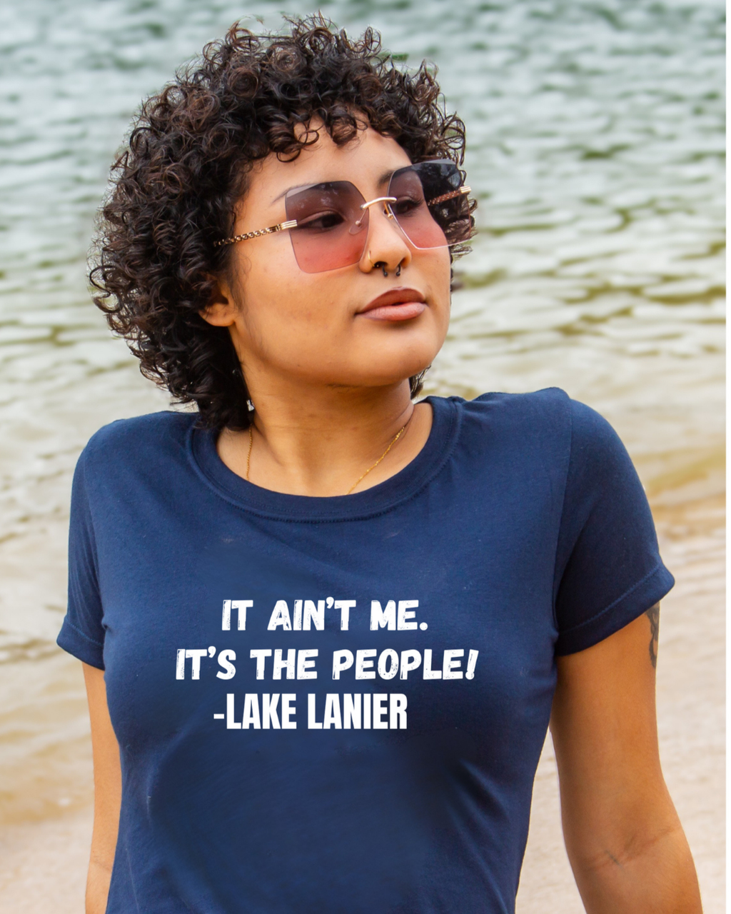 A woman with curly dark hair and pink-tinted sunglasses stands outdoors near the water, wearing a navy blue T-shirt with the white printed text 'IT AIN'T ME. IT'S THE PEOPLE! -LAKE LANIER'.